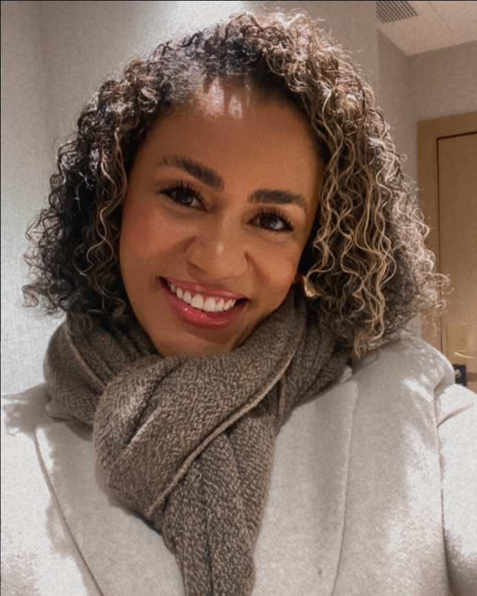 Psicóloga Adriana Andrade sorrindo, com cabelos cacheados e cachecol, em um ambiente acolhedor